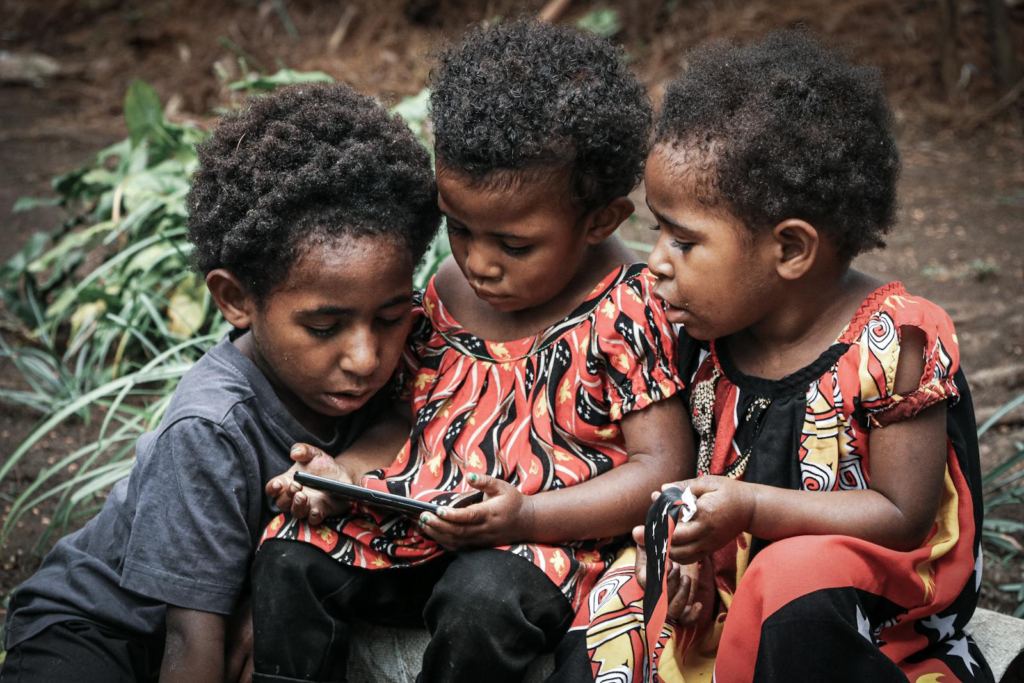 Three children using a smartphone outdoors in Papua New Guinea, exploring technology together.